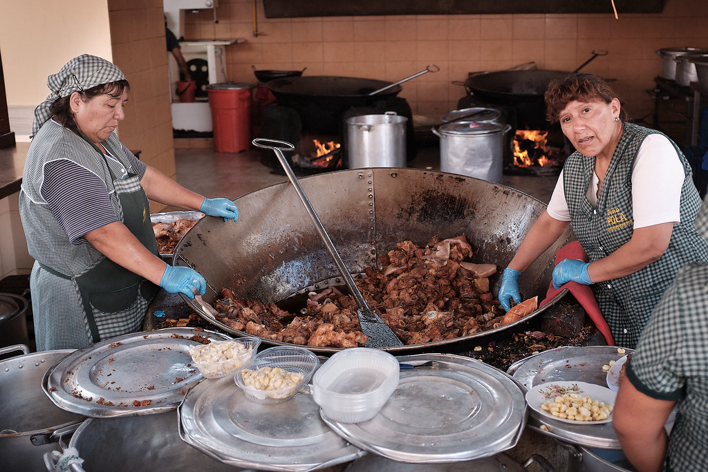 Cocineras de Doña Pola sirviendo chicharrón desde un perol gigante, con el fuego a leña encendido al fondo