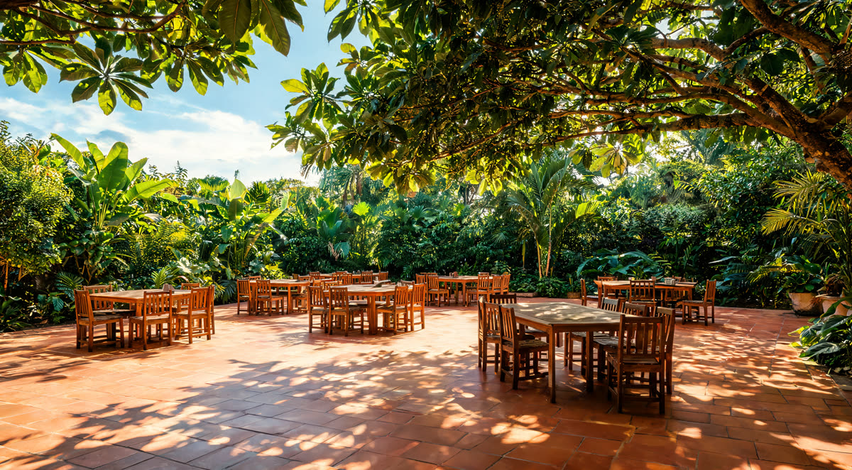 Patio tropical con mesas de madera bajo árboles frondosos y piso de terracota